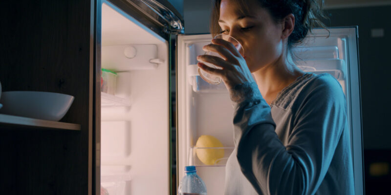 Thirsty woman standing in front of the fridge and drinking water late at night